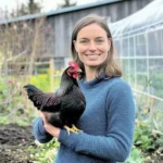 Eine lächelnde Frau hält ein Huhn in den Armen, während sie in einem Garten steht.